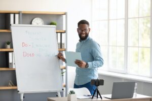 Happy young black man teacher in glasses looks at phone webcam shows to blackboard, shoots lesson